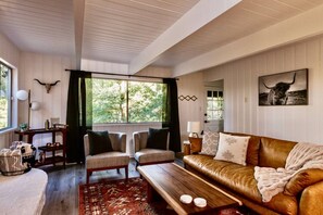 Un salon chaleureux avec un canapé en cuir, deux fauteuils, une table basse en bois et une grande fenêtre agrémentée de rideaux.