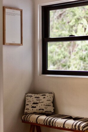 Una habitación de esquina con ventana, un cuadro en la pared y un banco con cojines estampados.