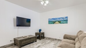 Un salon comprenant un téléviseur à écran plat, une table basse en bois, un canapé beige, une petite table d’appoint avec un vase bleu et un tableau représentant une scène de plage.