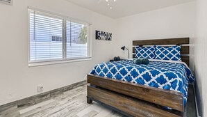 Une chambre à coucher comprenant un lit, un ventilateur de plafond, une fenêtre avec des stores, un climatiseur fixé au mur et une petite œuvre d’art.
