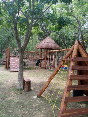 A playground with a rope net, wooden climbing structures, and a thatched-roof shelter.