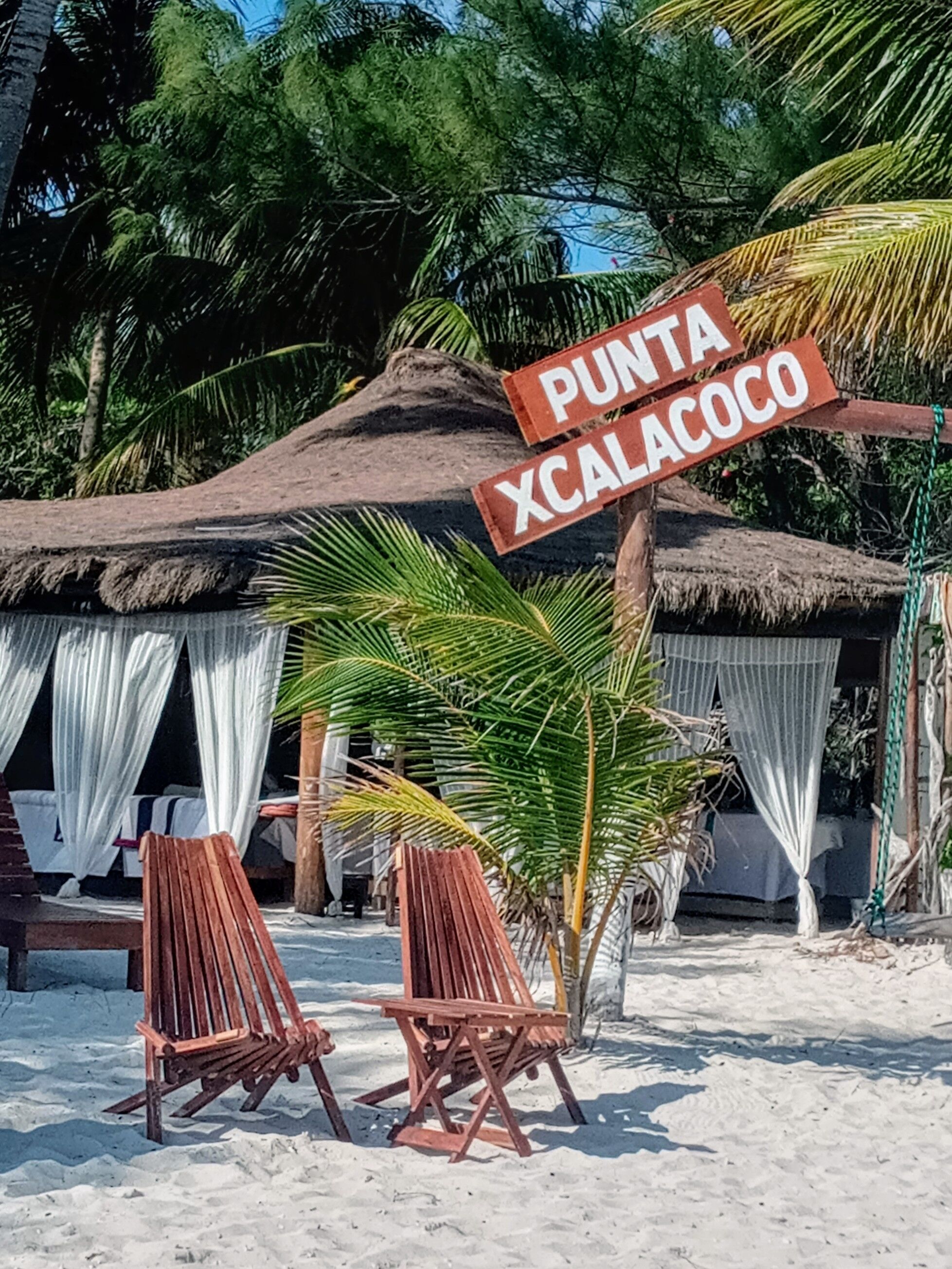 A beach setting with a sign reading "Punta Xcalacocho". There are two wooden chairs and a thatched-roof structure with white curtains.