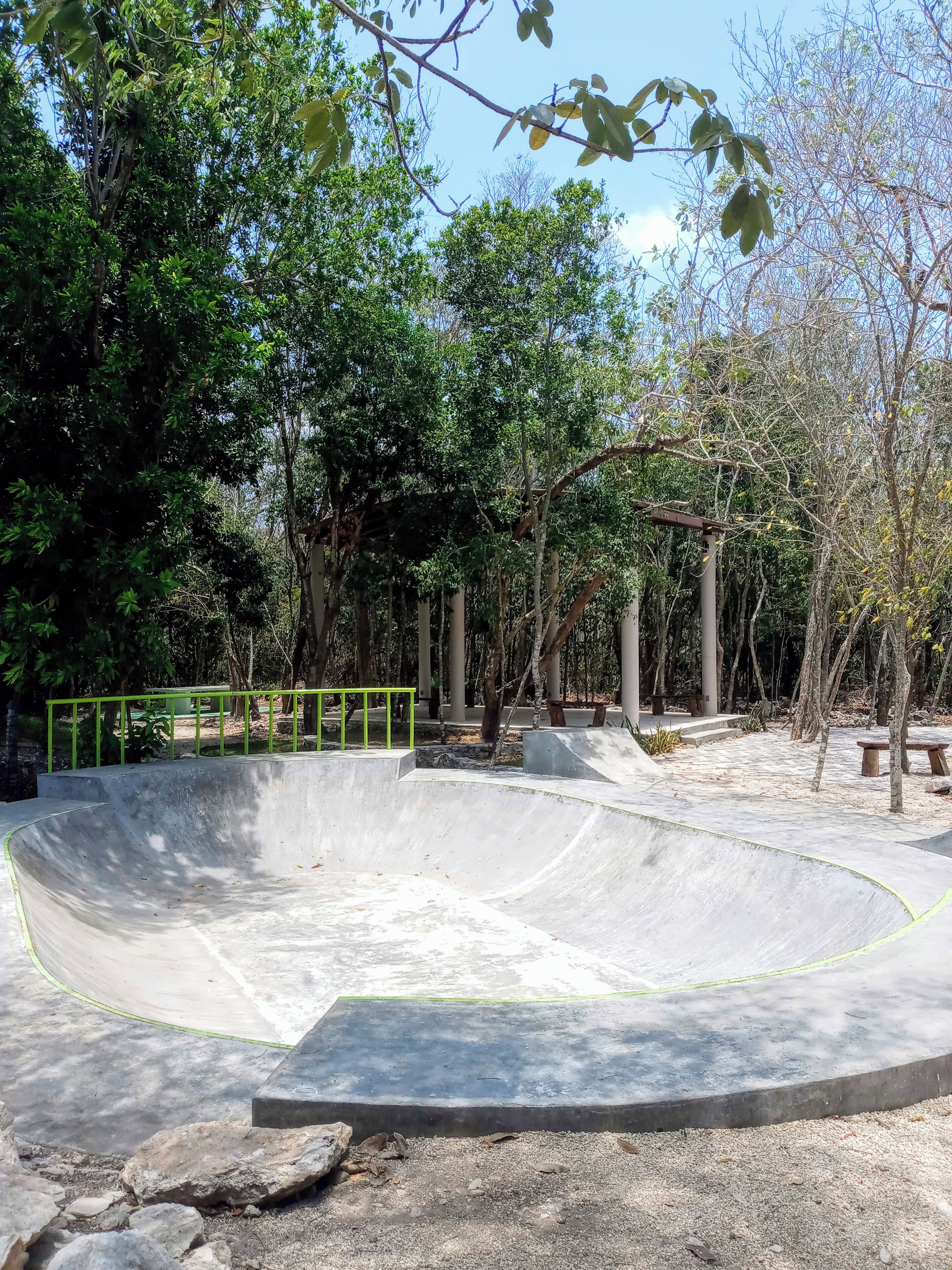 Een betonnen skatepark met een gebogen helling en een vlak gedeelte, omgeven door bomen en een groene hekken.