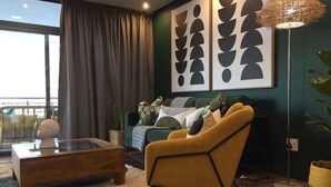 Ein modernes Wohnzimmer mit einem Glastisch, einem gelben Sessel und einem grünen Sofa mit gemusterten Kissen. An den Wänden befinden sich abstrakte Kunstwerke und eine hohe Stehlampe.