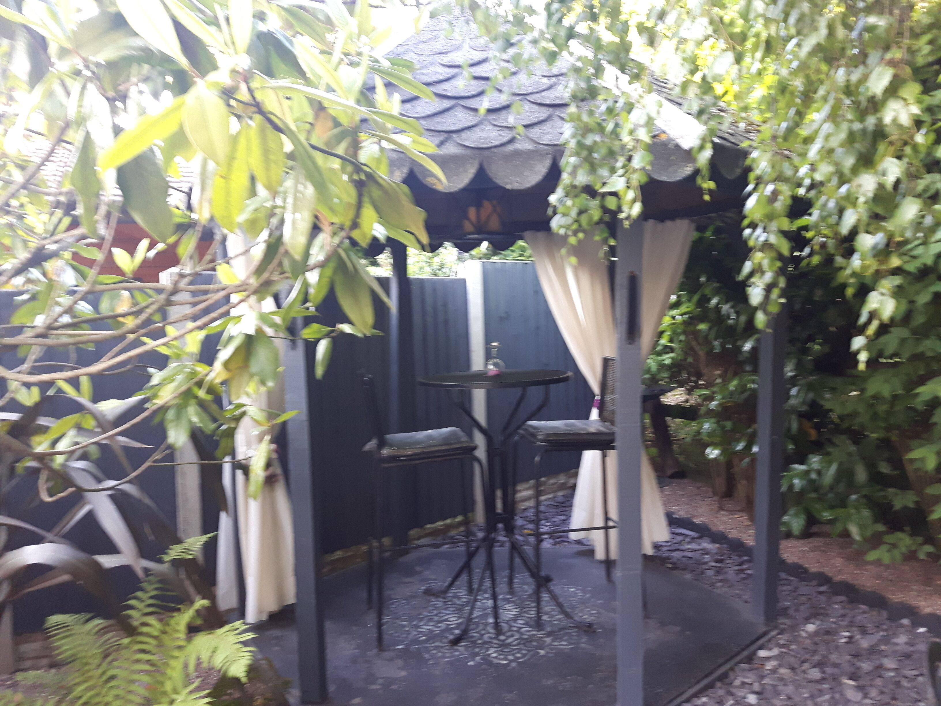 A covered outdoor seating area with two chairs and a table, surrounded by greenery and a decorative fence.