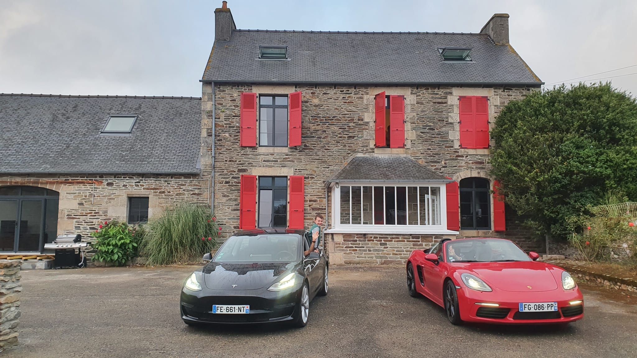 Een stenen huis met rode luiken, twee geparkeerde auto's en een tuin.