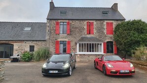 Una casa de piedra con contraventanas rojas, dos coches aparcados y un jardín.