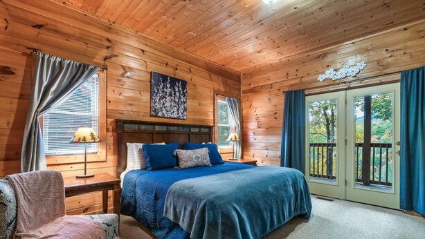 A wooden cabin bedroom with a bed, a chair, a nightstand, and a window with curtains. - Fall Family Fun l Views, Hot Tub, Theater & Pool l 2 King Beds (Gatlinburg)