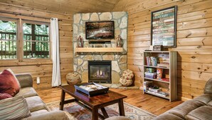 A cozy cabin interior with a stone fireplace, wooden walls, and a bookshelf. - Fall Family Fun l Views, Hot Tub, Theater & Pool l 2 King Beds (Gatlinburg)