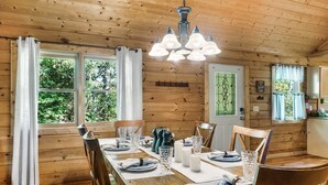 A wooden cabin dining area with a table set for four, surrounded by chairs, a chandelier, and windows with curtains. - Fall Family Fun l Views, Hot Tub, Theater & Pool l 2 King Beds (Gatlinburg)