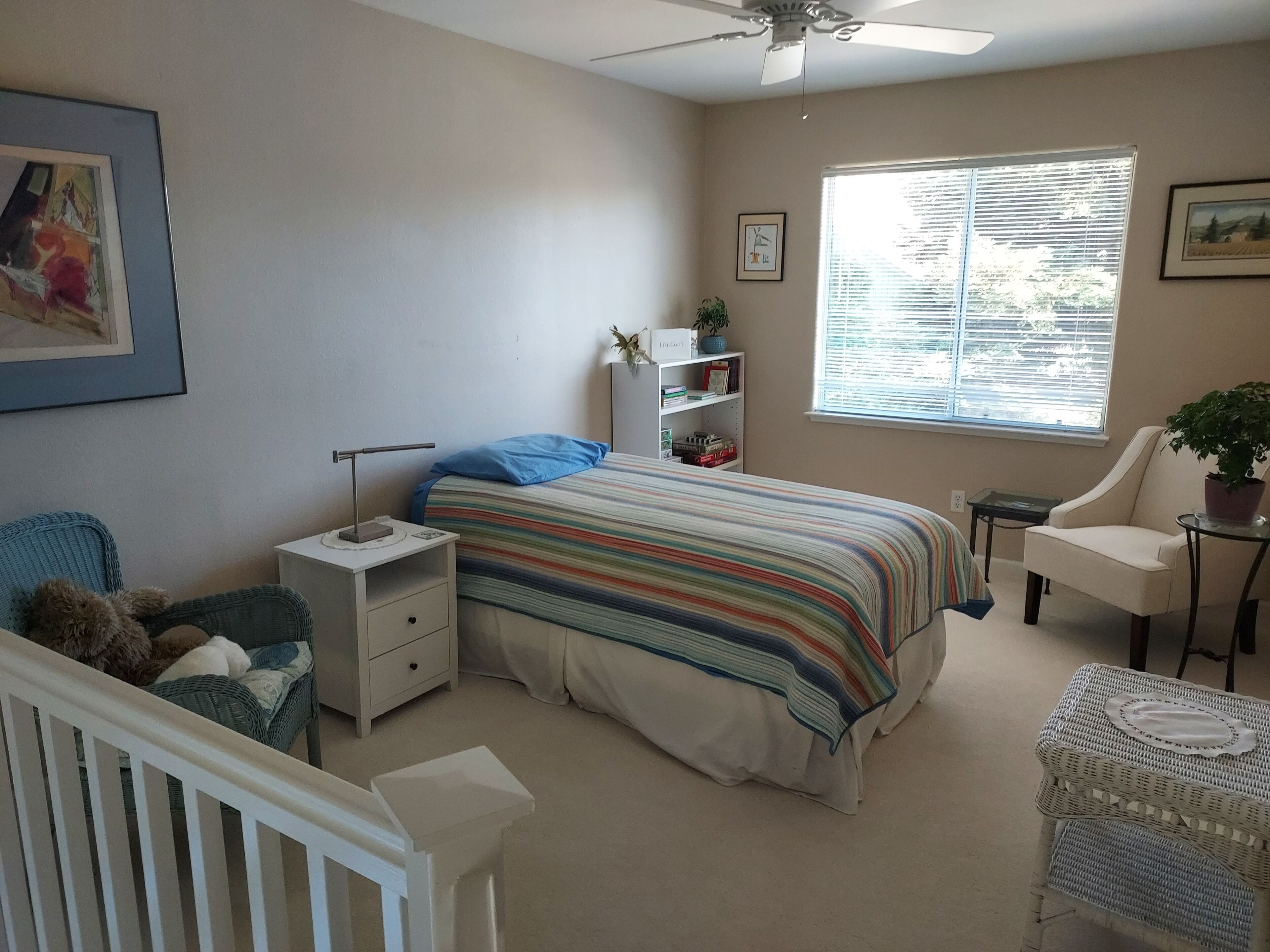 A bedroom with a bed, bedside table, nightstand, chair, window, and ceiling fan.