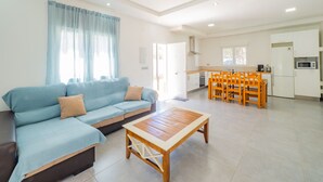 Una sala moderna con sofá, una mesa de centro de madera y un comedor con mesa y sillas. Se ve una cocina con electrodomésticos al fondo.
