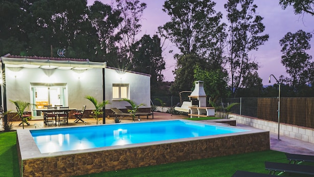 Una piscina con borde de piedra, zona de barbacoa y patio con sillas y una mesa.