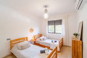 Una habitación con dos camas de madera, una mesita con lámpara, una ventana con cortinas y un aire acondicionado.