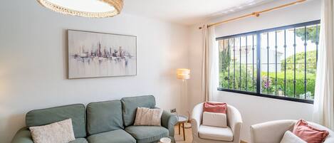 Ein modernes Wohnzimmer mit einem grünen Sofa, einem weißen Sessel, einem runden Couchtisch und einem großen Fenster mit Blick ins Grüne.