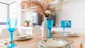 Ein Esstisch ist gedeckt mit blauem Glas, blumigen Tellern und einer weißen Vase mit Trockenpflanzen.