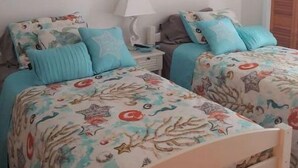 Une chambre avec deux lits, chacun agrémenté de draps à motifs floraux et d’oreillers assortis. Deux tableaux encadrés ornent le mur, ainsi qu’une table de chevet avec une lampe et une porte en bois.