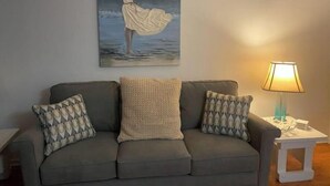 Un salon avec un canapé gris, des coussins à motifs, un tableau représentant une femme au bord de la mer et une lampe de table.