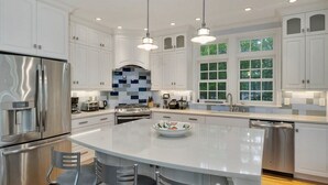 A modern kitchen with white cabinets, stainless steel appliances, a large island with bar stools, and a view of greenery outside.