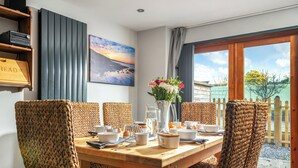 A dining area with a wooden table set for four, surrounded by woven chairs, a vase with flowers, and a painting on the wall. - Coach House - UK38039 (Honeyborough)