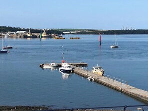 A harbor with boats docked at a pier, a lighthouse in the distance, and buildings on the shore.