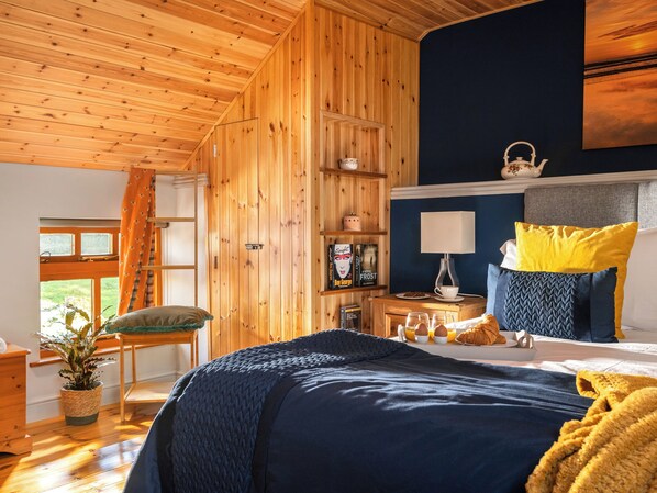 A cozy bedroom with a wooden ceiling, a bed with a blue and yellow pillow, a bedside table with a lamp, and a bookshelf with books and decorative items.