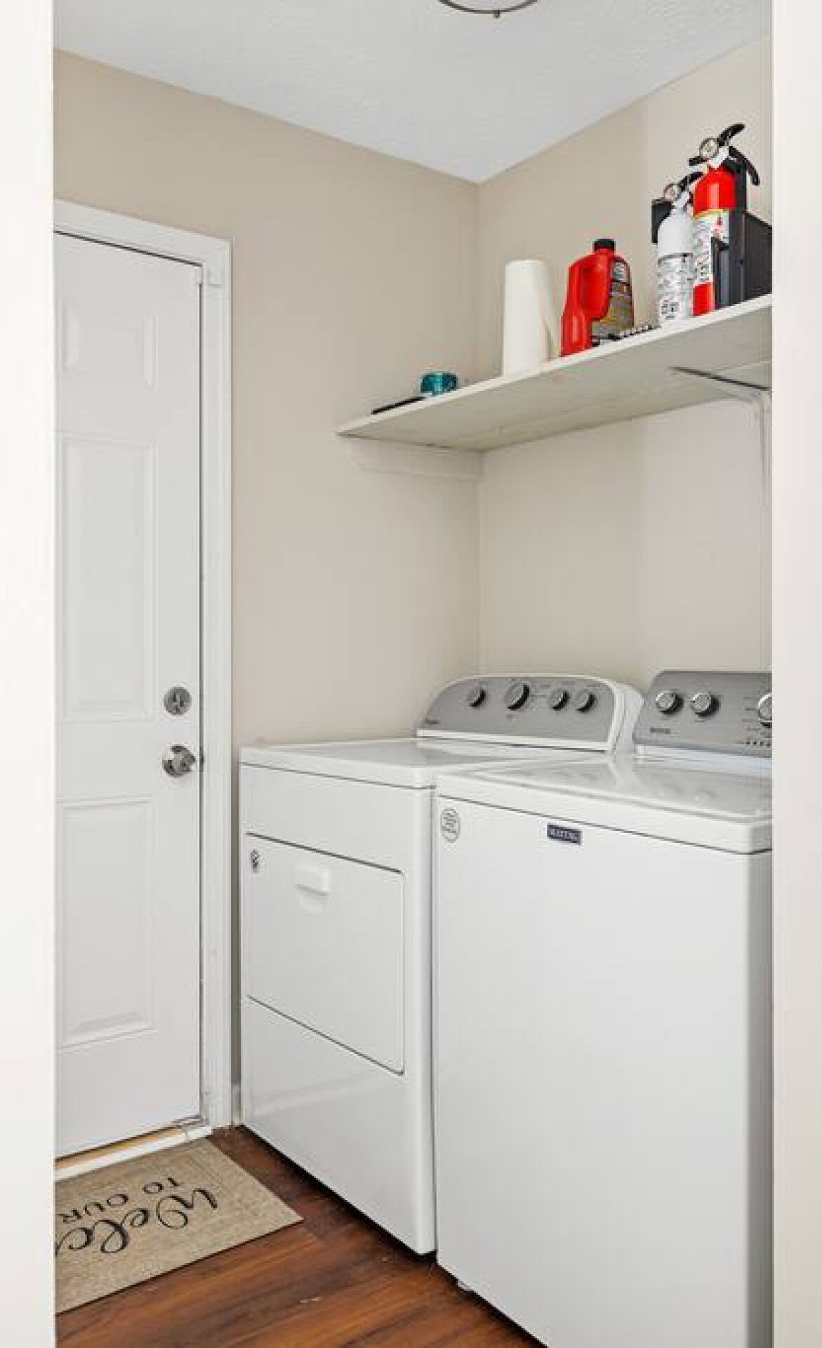 Une buanderie avec un lave-linge et un sèche-linge blancs, une porte blanche et une étagère au-dessus où sont rangés les produits de nettoyage.