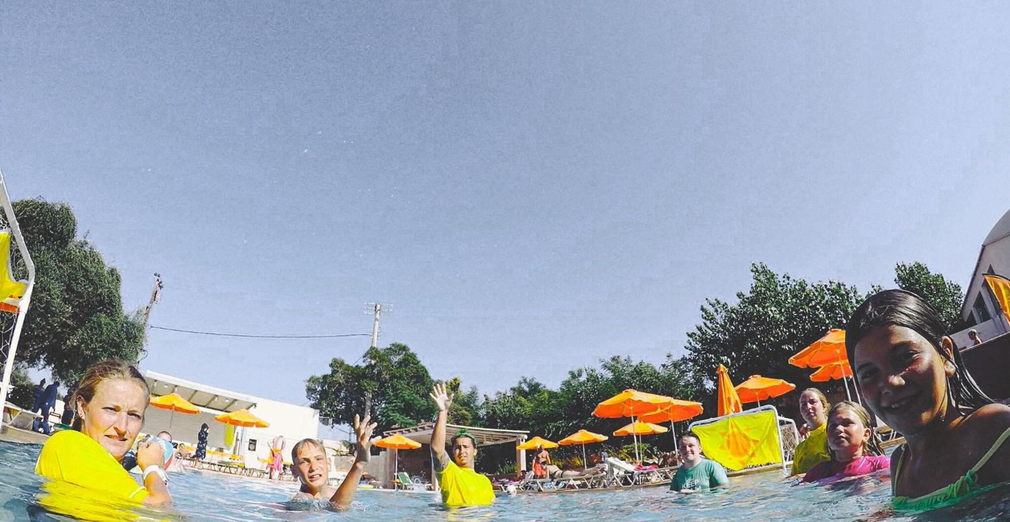 A swimming pool area with multiple people in the water, surrounded by umbrellas and lounge chairs.
