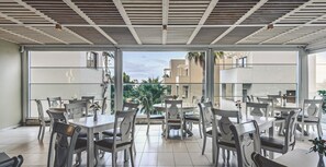 A dining area with tables and chairs, large windows, and a view of buildings and greenery.