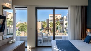 A hotel room with a bed, a TV, a desk, and a balcony with a view of buildings and palm trees. - Althea Village Hotel (Chania)
