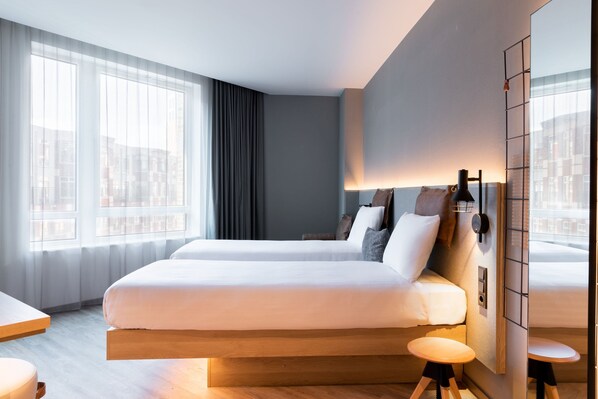 A modern hotel room with a large bed, bedside tables, and a view of the city through large windows.