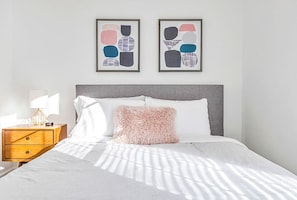 Uma cama bem arrumada com um travesseiro fofo, uma mesa de cabeceira com abajur, e duas obras de arte abstratas nas paredes.