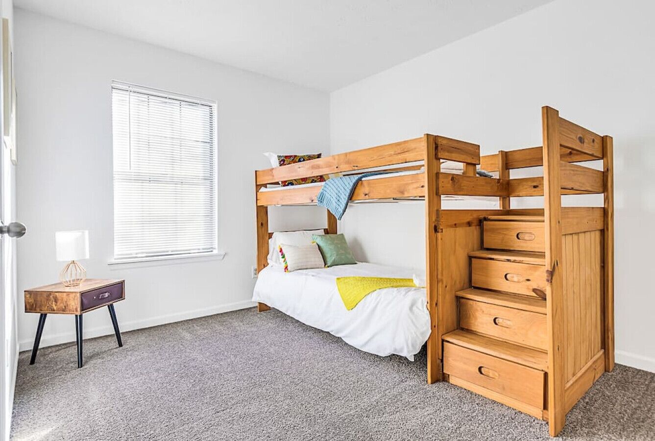 A bunk bed with a wooden ladder and drawers, a small bedside table, and a window with blinds.
