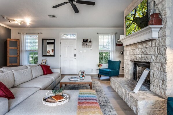 A living room with a stone fireplace, a ceiling fan, and a large window with curtains.