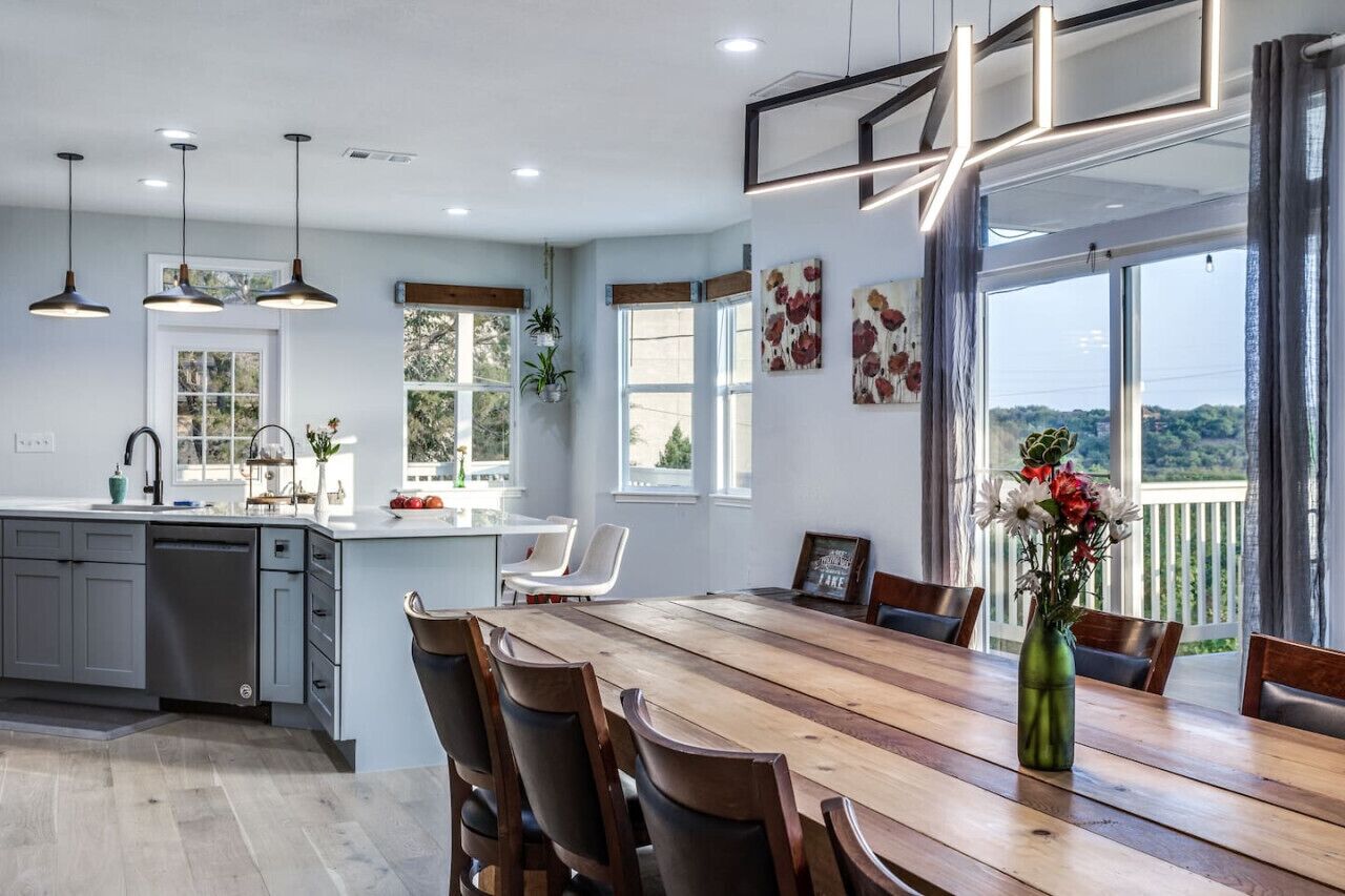 Une cuisine moderne avec une table à manger en bois, des armoires grises, des luminaires suspendus et de grandes fenêtres offrant une vue sur l’extérieur.