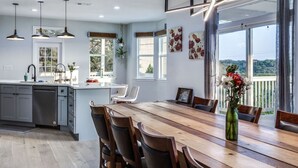 Uma cozinha moderna com mesa de jantar de madeira, armários cinzas, luminárias pendentes e grandes janelas com vista para o exterior.