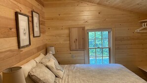 Ein Schlafzimmer einer Holzhütte mit Bett, Kissen, Nachttisch, Lampe, Kleiderschrank und einem Fenster mit Blick auf Grün.