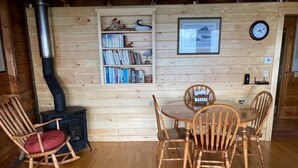 Ein gemütliches Holzhaus-Interieur mit Esstisch und Stühlen, einem Bücherregal, einer Uhr und einem Kaminofen.
