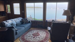 Ein gemütliches Wohnzimmer mit einem blauen Sofa, einem gemusterten Teppich und Meerblick durch große Fenster.