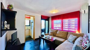 A compact living space with a sofa, a coffee table, and a TV. There's a kitchen area visible through an open door.