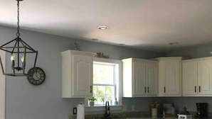 A modern kitchen with white cabinets, a dining table with chairs, a hanging light fixture, and a window providing natural light.