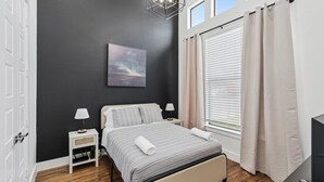 Un dormitorio con cama, mesitas de noche, lámpara, un cuadro, ventana con cortinas y televisión.