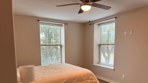 Ein Schlafzimmer mit einem Bett, einem Nachttisch, einer Lampe und einem Deckenventilator. Es gibt zwei Fenster mit Jalousien.