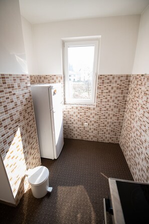 Cocina pequeña y estrecha con un refrigerador blanco, un inodoro blanco y una ventana que proporciona luz natural. Las paredes están cubiertas de azulejos con un diseño marrón y blanco.