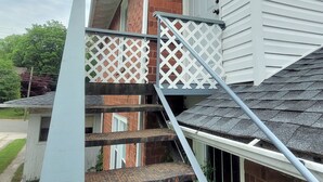 A residential staircase with a metal handrail and a white lattice guardrail, leading to a brick house with white siding. - Vintage One Bedroom with Living/Kitchen (Mount Airy)
