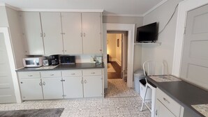 A compact kitchen with white cabinets, a microwave, a toaster, and a coffee maker. There's a small table with a chair, a TV mounted on the wall, and a hallway leading to another room. - Vintage One Bedroom with Living/Kitchen (Mount Airy)