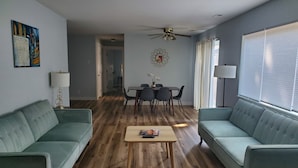 Een woonkamer met een houten vloer, twee groene banken, een salontafel, een eettafel met stoelen, een plafondventilator en een schilderij aan de muur.