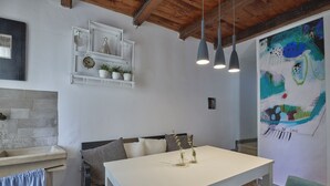 A modern kitchen with white cabinetry, a large white table, and pendant lights. There is a shelf with plants and a clock, and a colorful abstract painting on the wall. - Valesi holiday home (Rakalj)
