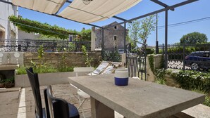 An outdoor patio area with a table and chairs, a hanging light fixture, and a view of a stone building and parked cars. - Valesi holiday home (Rakalj)