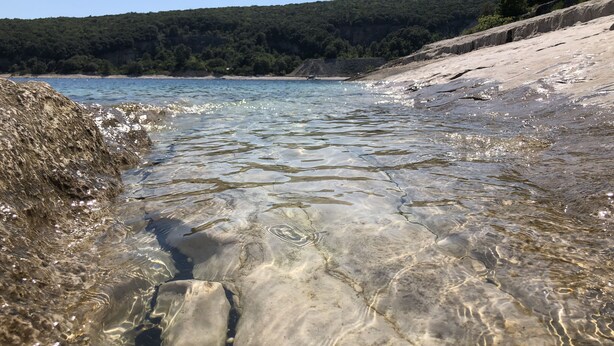 A clear view of a rocky shoreline with calm water, a sandy beach, and a forested hill in the background. - Valesi holiday home (Rakalj)